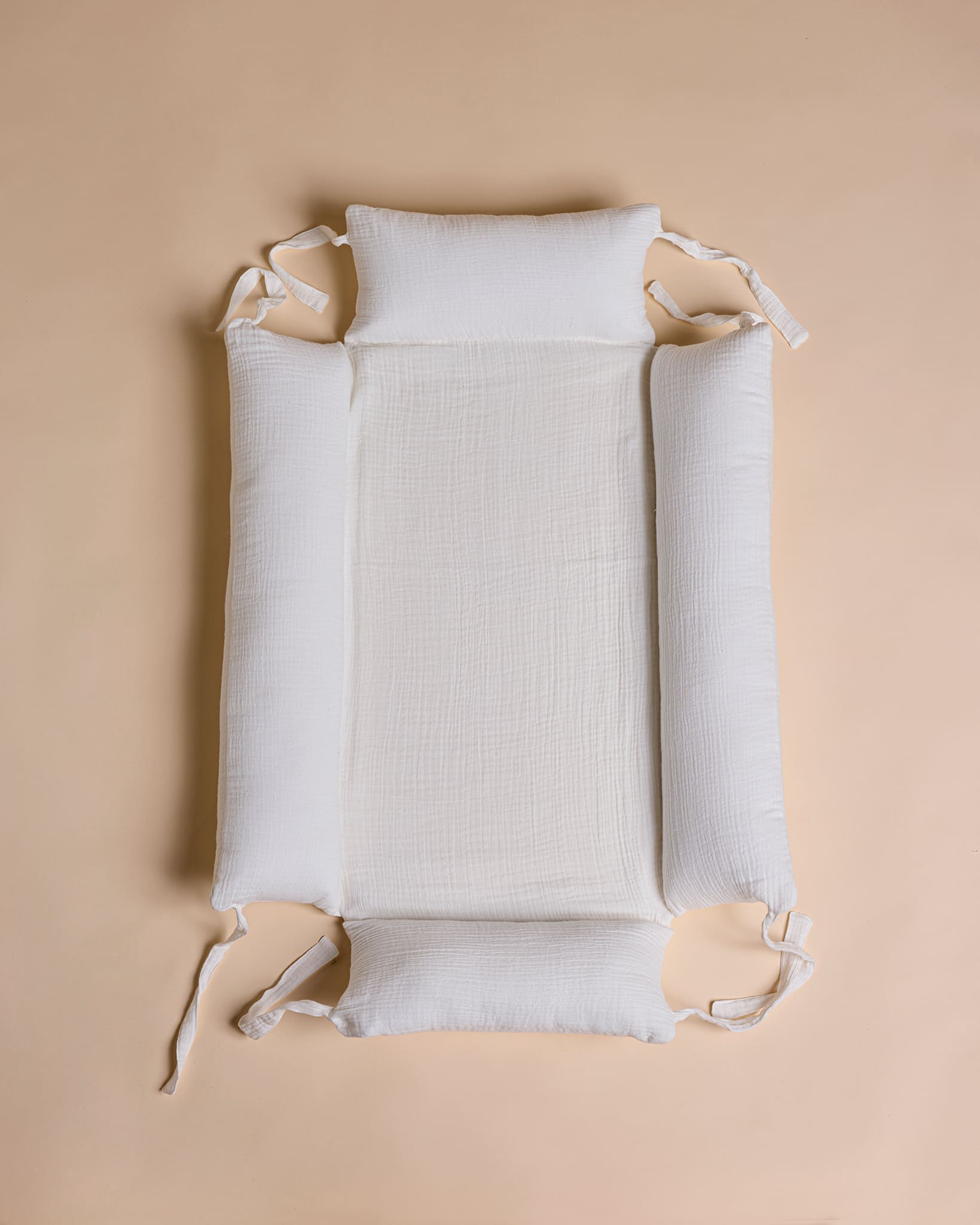 A white textured baby mattress guard with integrated side bolsters and two small matching head and foot pillows, laid flat on a solid beige background.