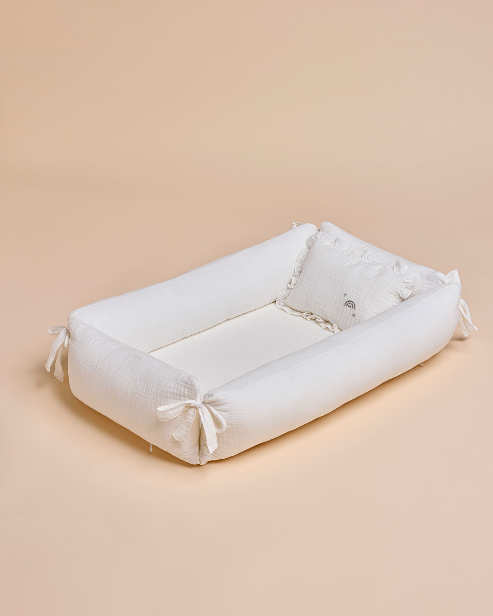 A white fabric baby changing pad with padded corners and tie straps, shown on a solid beige background.